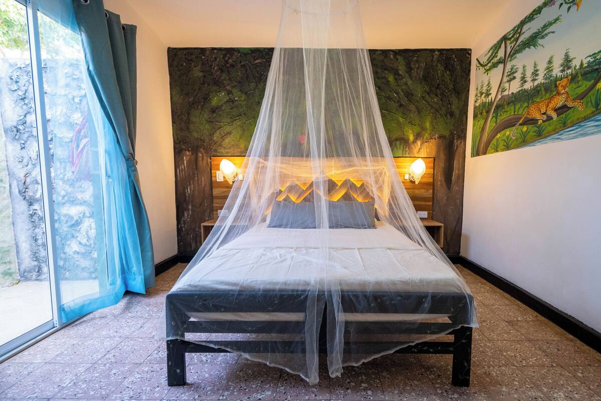 Una cama con dosel, una mesita de noche, una pared con un cuadro y una ventana con cortinas.
