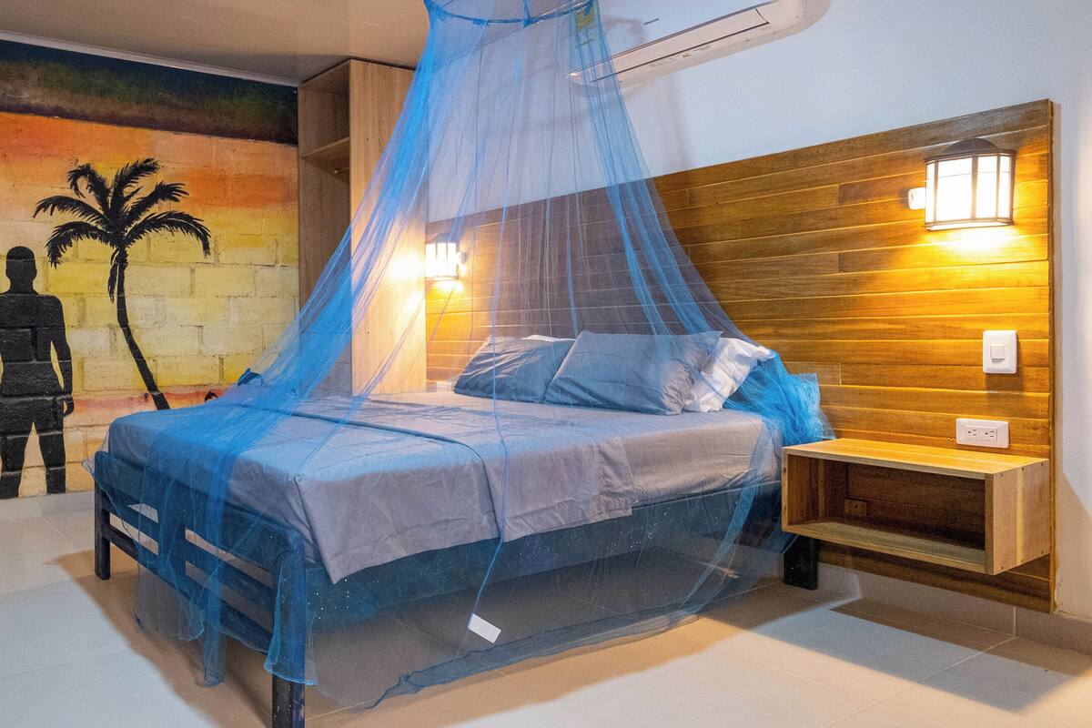 Un dormitorio con una cama bajo una mosquitera azul, un cabecero de madera, una mesita de noche y un mural en la pared.