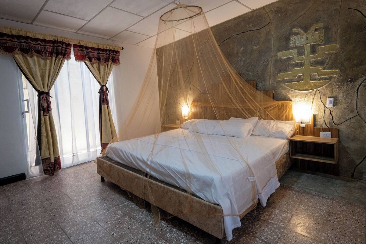 Un dormitorio con cama, cortinas, una mesita de noche con lámpara y una pared decorada con un emblema.
