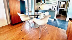Een moderne keuken met een eethoek, met een glazen tafel en witte stoelen, houten vloeren en uitzicht door een groot raam.