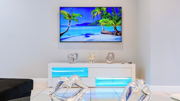 Un soggiorno moderno con una TV a schermo piatto montata a parete che mostra una spiaggia tropicale. Davanti alla TV c'è una consolle bianca con un dettaglio blu, e due sculture astratte trasparenti sul pavimento.
