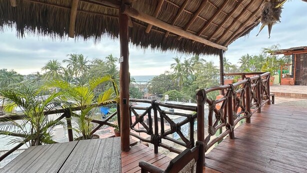 A wooden deck with a thatched roof, a wooden railing, and a view of palm trees and a body of water. - Habitación Lupe Gil. (San Francisco)