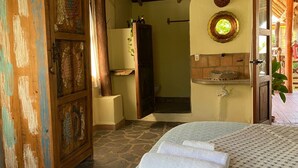 A room with a stone floor, a bed with white linens, a wooden door with carvings, and a shower area. - Habitación Lupe Gil. (San Francisco)