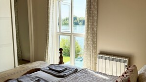 A bedroom with a large bed, a window with curtains, a wooden ceiling, and a view of greenery outside. - Peaceful Lakefront Escape w/ Sunset Views (South Cerney)