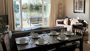 A dining area with a table set for four, a sofa, and a view of a lake through large windows. - Peaceful Lakefront Escape w/ Sunset Views (South Cerney)