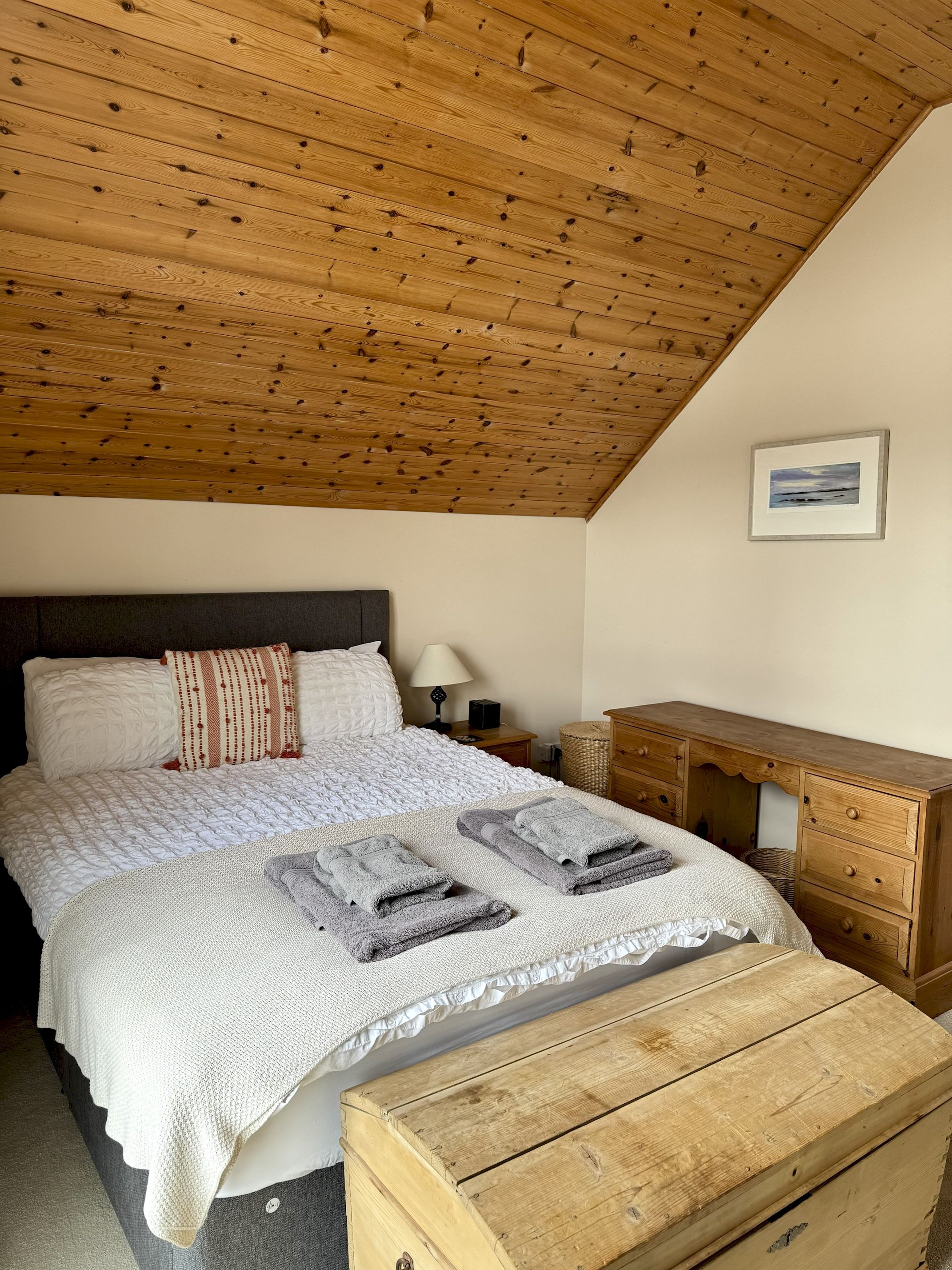 Een slaapkamer met een houten plafond, een bed met wit beddengoed, een nachtkastje, een dressoir en een ingelijste foto aan de muur.
