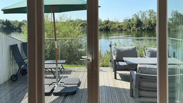 A deck with outdoor furniture and a green umbrella, overlooking a lake. - Peaceful Lakefront Escape w/ Sunset Views (South Cerney)