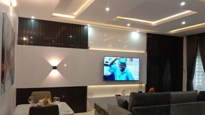 A modern living room with a flat-screen TV mounted on the wall, a dining area with a table and chairs, and a grey sofa.
