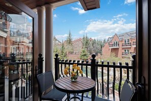A balcony with a round table and chairs, a vase with yellow flowers, and a view of brick buildings and trees. - The Aspen Mountain Residences 24A - Studio Residence (Aspen)