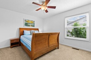 Une chambre à coucher avec un lit en bois, une table de chevet, un ventilateur de plafond, une fenêtre avec des stores et un tableau accroché au mur.