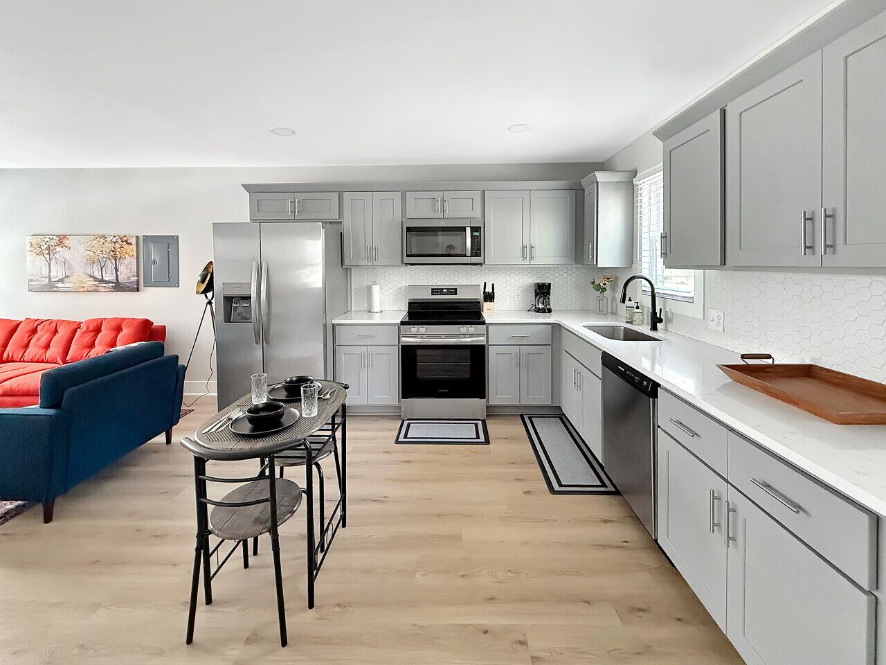 Eine moderne Küche mit grauen Schränken, einem Edelstahlkühlschrank, einer Mikrowelle und einem eingebauten Backofen. Ein Essbereich mit Tisch und Stühlen sowie eine rote Couch im Hintergrund sind ebenfalls vorhanden.