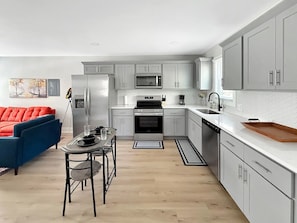 A modern kitchen with grey cabinets, a stainless steel refrigerator, a microwave, and a built-in oven. There is a dining area with a table and chairs, and a red sofa in the background.