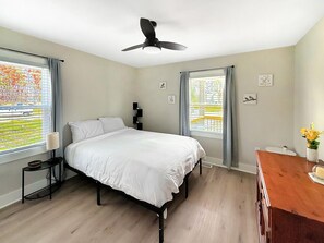 A bedroom with a bed, a ceiling fan, a nightstand, a lamp, a window with curtains, and a view of the outside. - 1BR Garner Gem|Backyard & Pet Friendly (Garner)