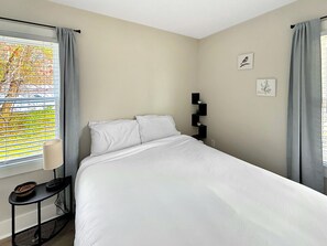 A neatly made bed in a room with a window, a small table with a lamp, and two wall-mounted shelves with decorative items.