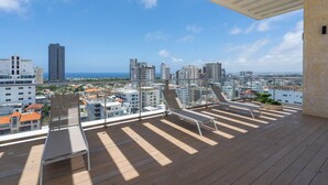 A balcony with two sun loungers and a view of a cityscape with buildings and the sea. - RELAXING*1BR*Pool |Gym* Rooftop *yellowkey* (Santo Domingo)