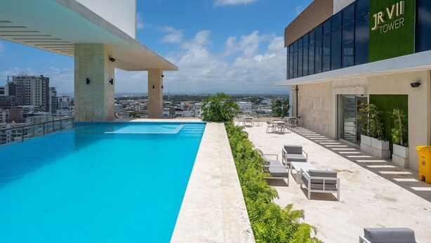 A rooftop pool with a clear view of the cityscape, a modern building with 'JR VII TOWER' signage, and outdoor seating area. - RELAXING*1BR*Pool |Gym* Rooftop *yellowkey* (Santo Domingo)