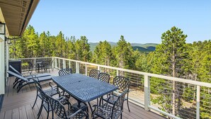 A covered outdoor deck with a table and chairs, surrounded by a white railing, overlooking a forested landscape with mountains in the distance. - Rocky Mountain Retreat - Hot Tub and EPIC views! (Rollinsville)