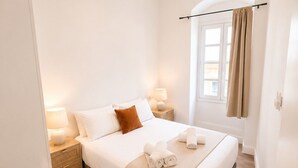 Un dormitorio ordenado con cama, lámparas de noche, una ventana con cortinas y suelo de madera.