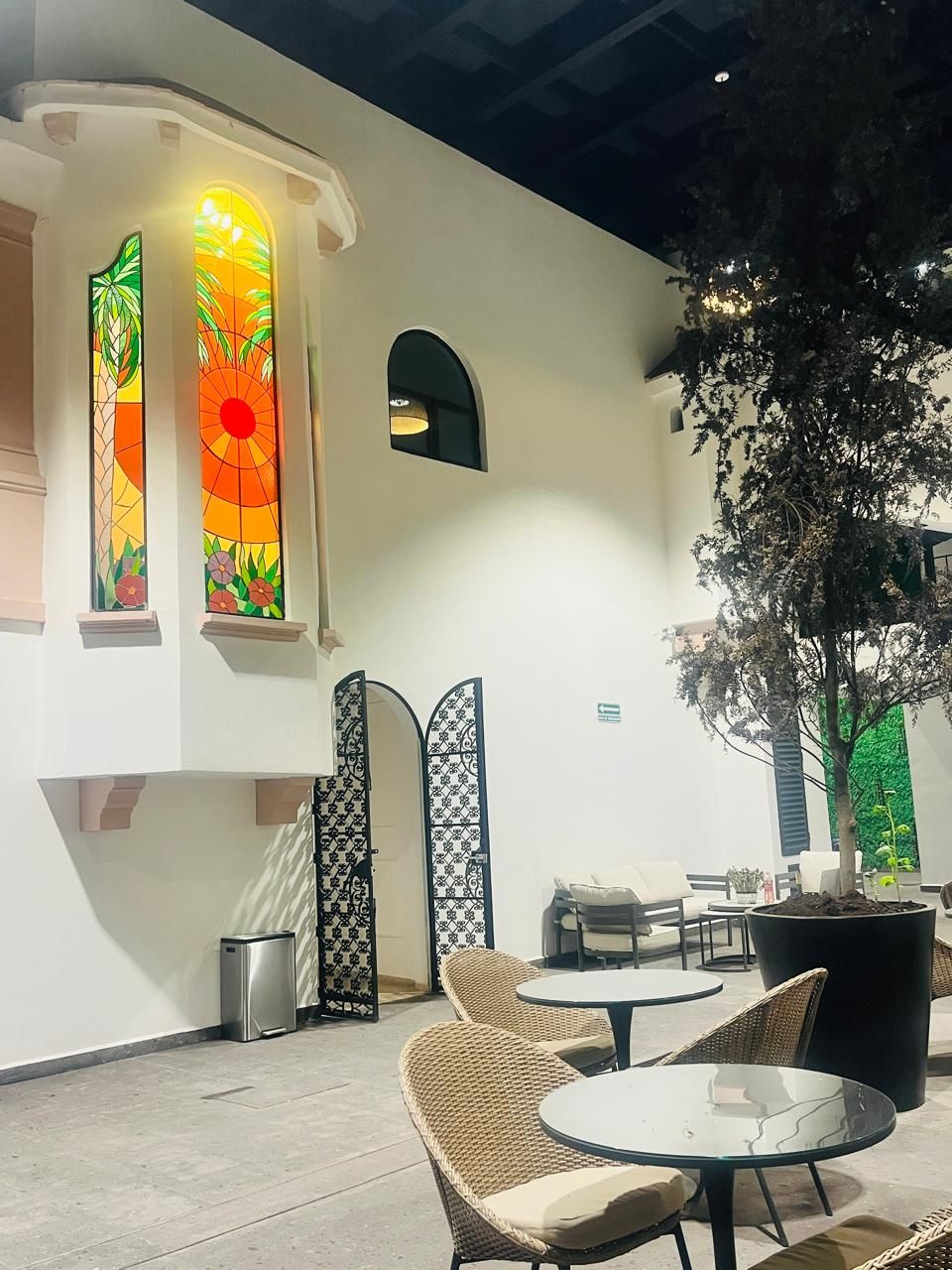 A modern outdoor seating area with wicker chairs and a glass table, a decorative tree, and a wall with stained glass windows.