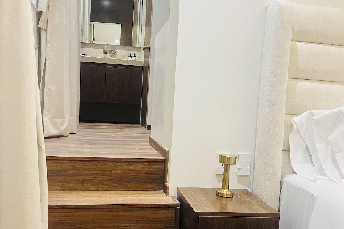 Une chambre d’hôtel avec un escalier en bois menant à la salle de bain, une table de chevet avec une lampe et un lit avec du linge de lit blanc.