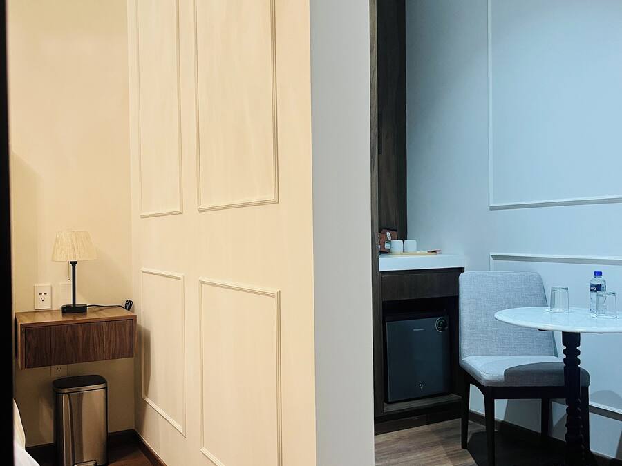 A hotel room with a wooden floor, a bed, a desk with a lamp, a trash bin, a small kitchenette, and a seating area with a table and chairs.