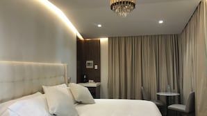 Ein Hotelzimmer mit einem großen Bett, einer Kristallleuchter, einem kleinen Tisch mit Stuhl und einem Fenster mit Vorhängen.