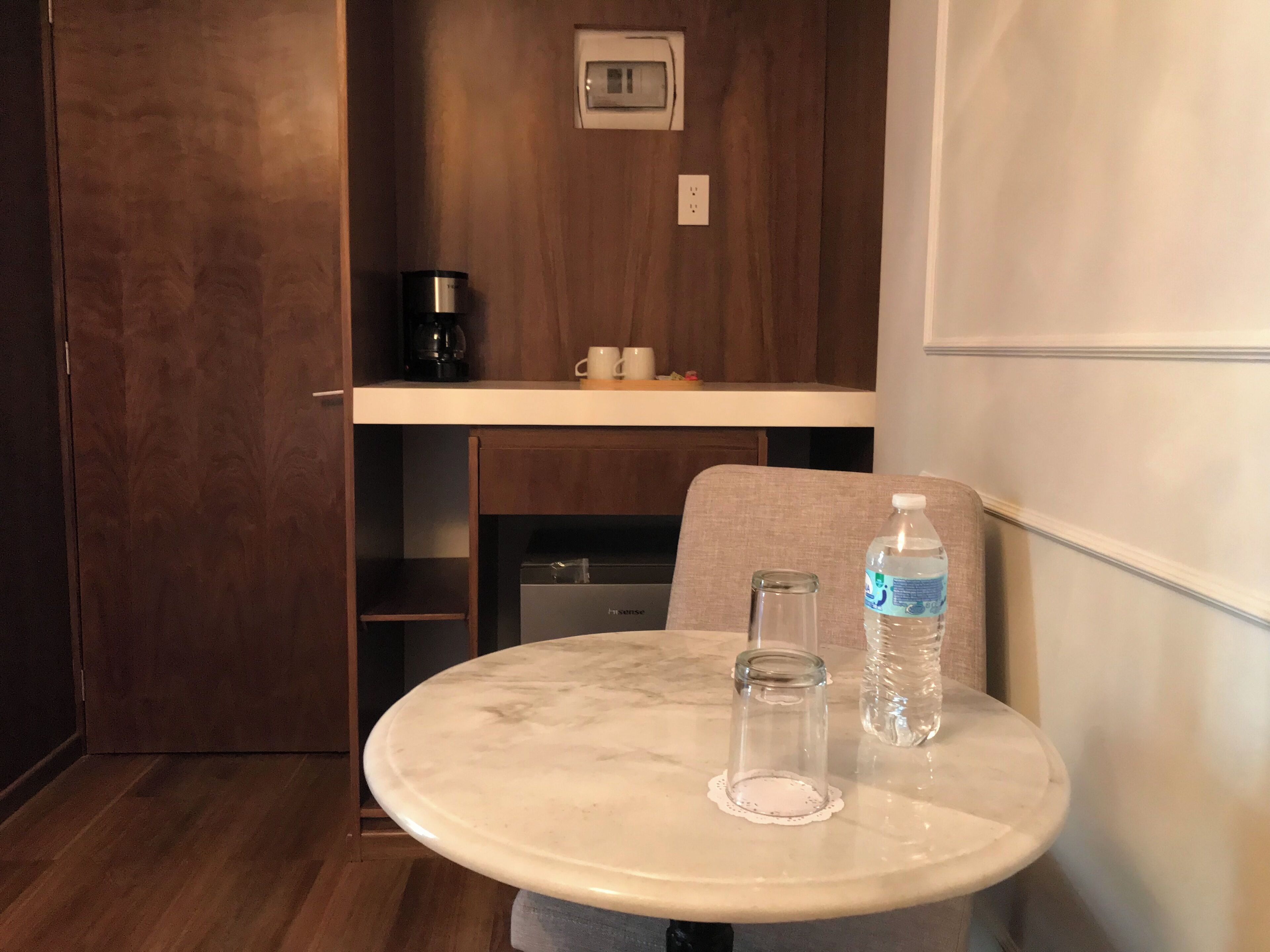 Ein kleiner, runder Tisch mit Marmorplatte, zwei klare Gläser und eine Wasserflasche. Darüber befindet sich ein Regal mit einer Kaffeemaschine und Tassen.