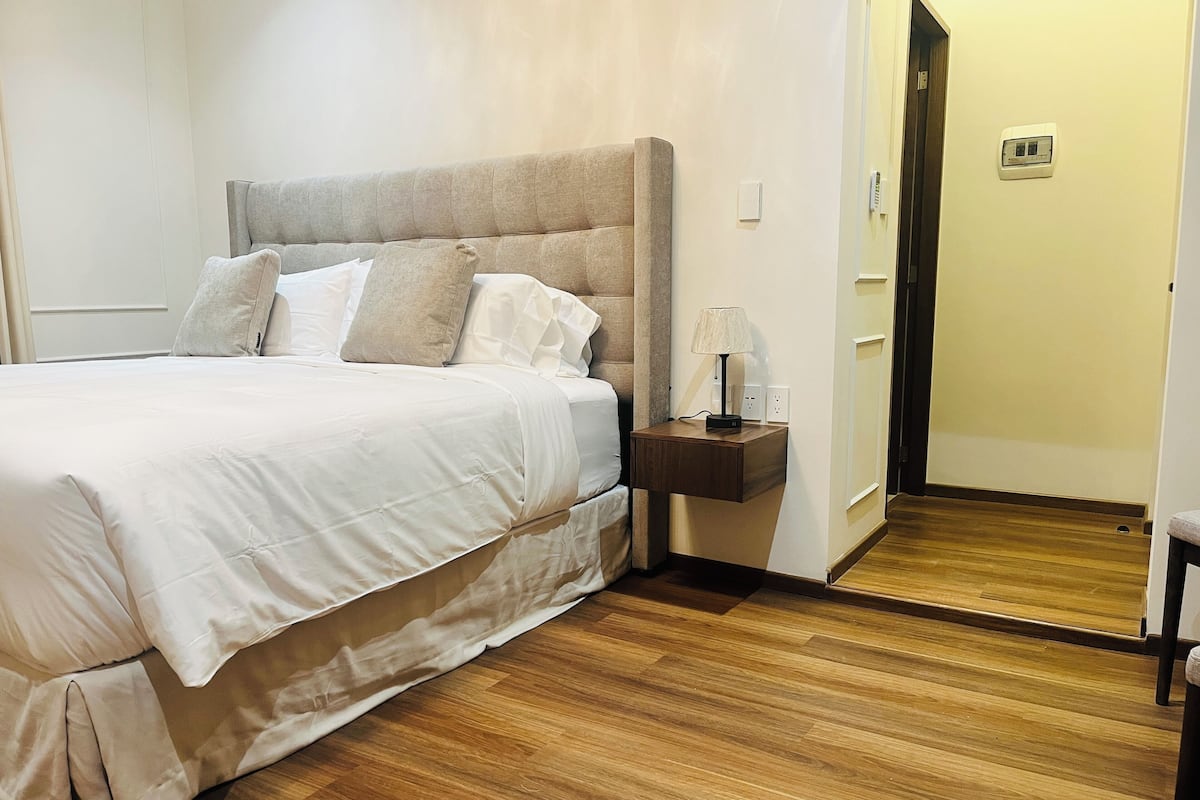 Un lit bien fait, avec du linge de lit blanc et une tête de lit en bois, une table de chevet avec une lampe et un plancher en bois.