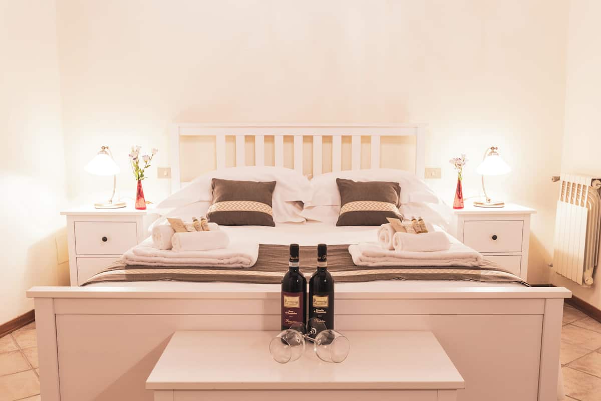 A neatly made bed with pillows, two bedside tables with lamps, a vase with flowers, and two bottles of wine on a bedside table.