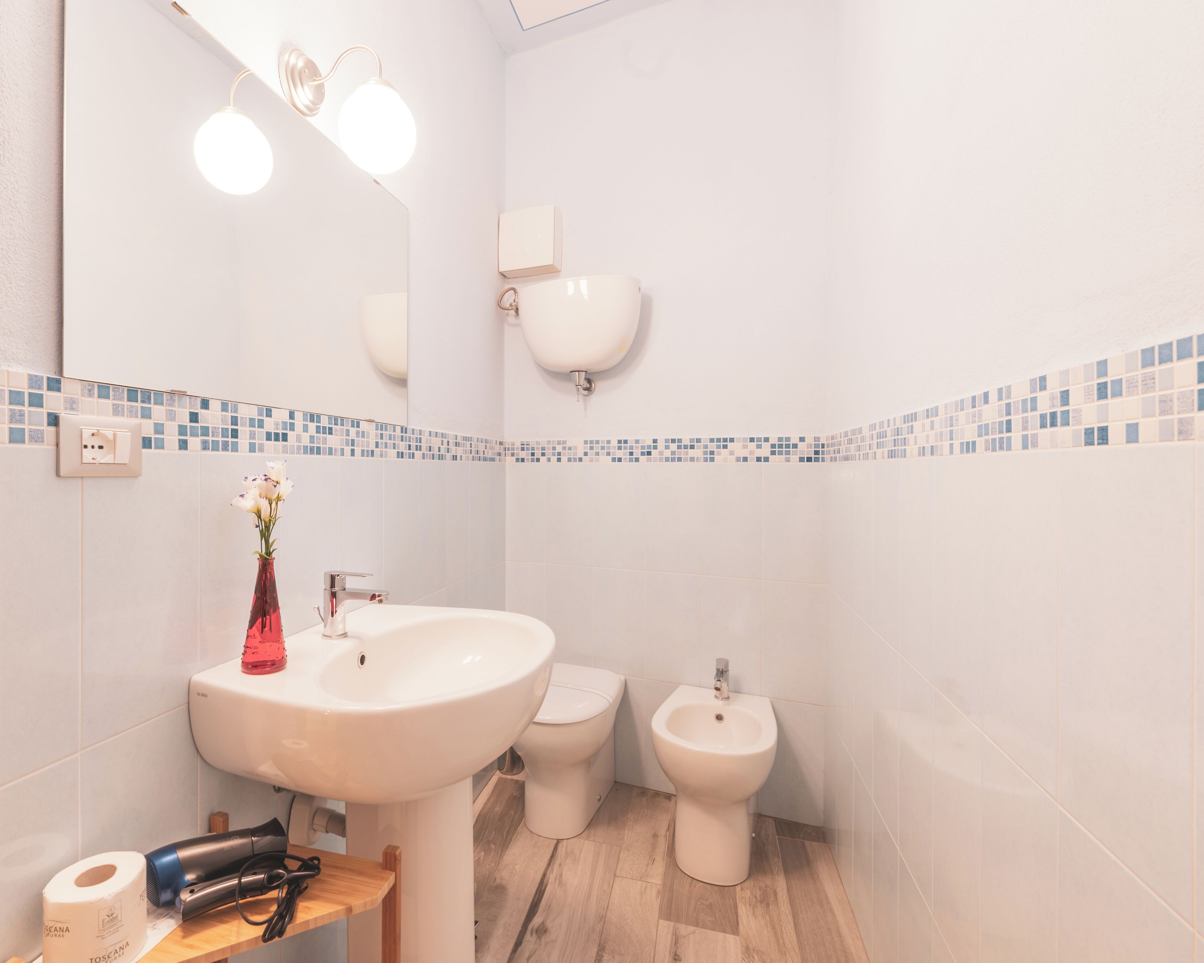 Een badkamer met een wastafel, spiegel, twee toiletten en een klein tafeltje met een haardroger en een rol toiletpapier.