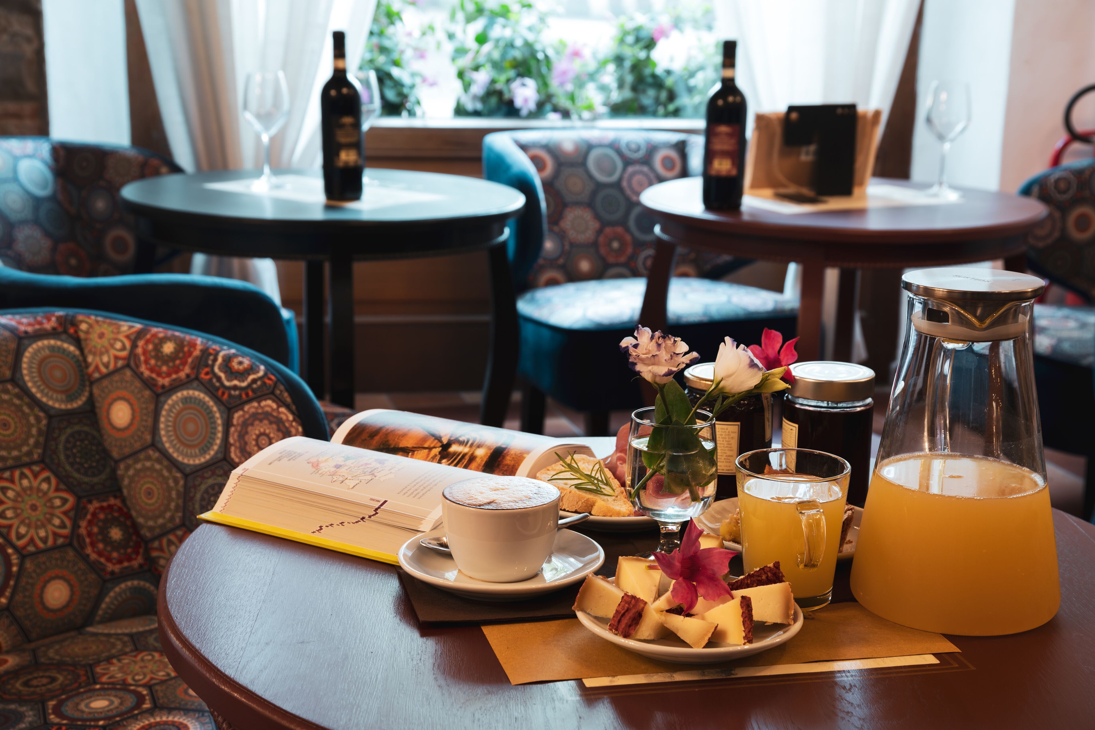 A dining table with a book, a cup of coffee, a glass of juice, a pitcher, and a plate of cheese and flowers.
