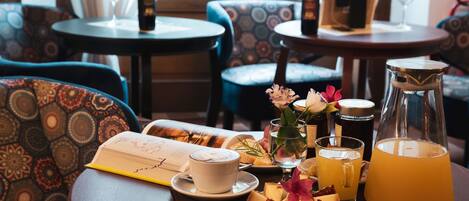 A dining table with a book, a cup of coffee, a glass of juice, a pitcher, and a plate of cheese and flowers.