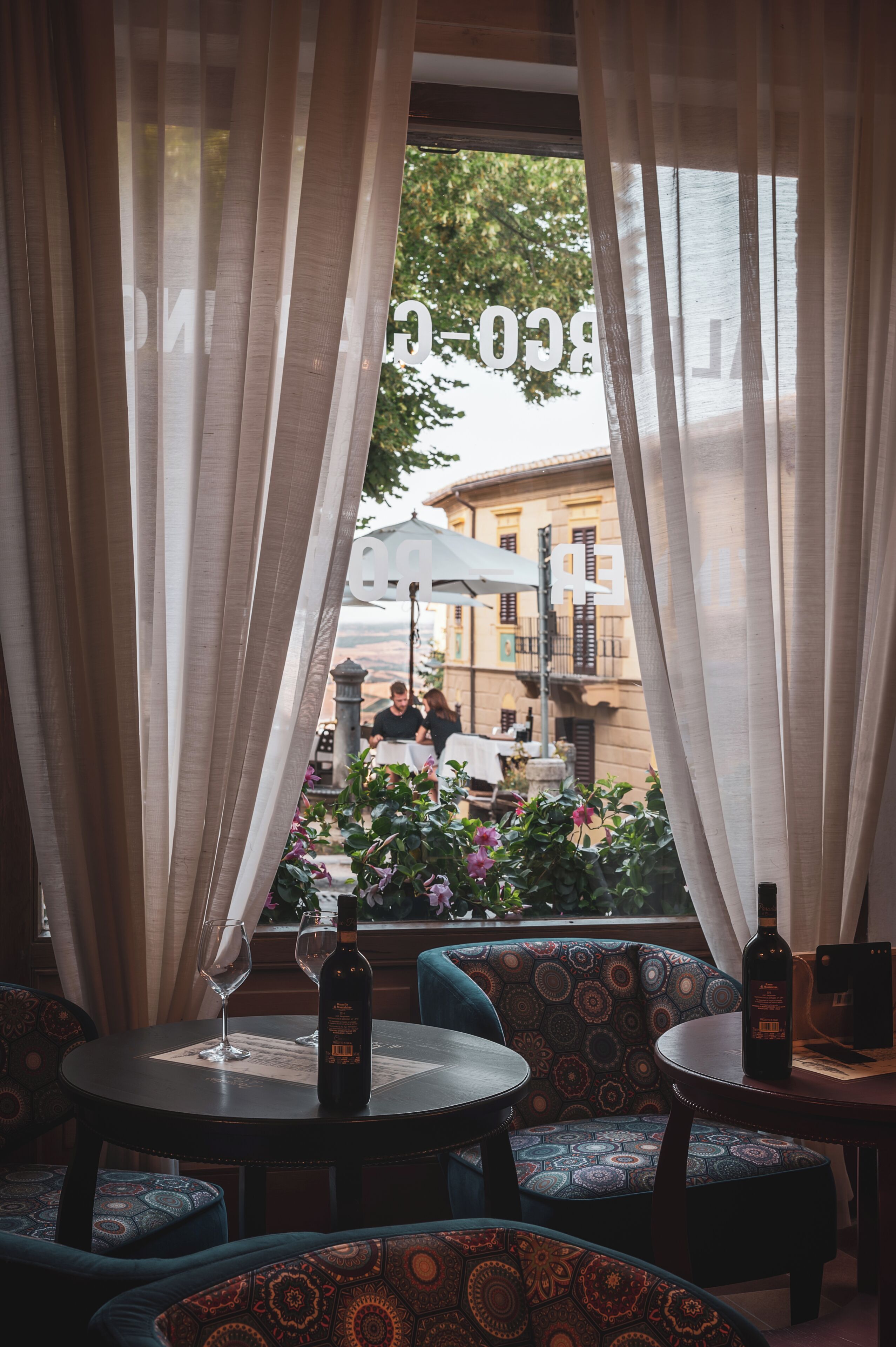 A restaurant with a view through sheer curtains, featuring round tables and patterned cushions.