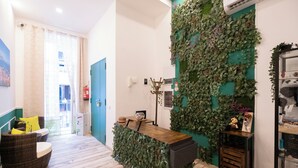 Un área de recepción con pared verde, mostrador de madera, sillas de mimbre y una pequeña estantería con objetos decorativos.