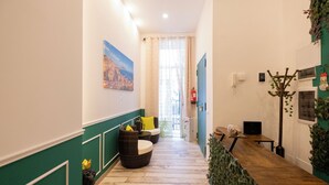 Un pasillo con suelo de madera, paredes verdes, mobiliario de mimbre y vistas a una habitación con ventana.