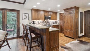 A kitchen with wooden cabinets, a central island with stools, a microwave, and a large window overlooking greenery. - The Charter at Beaver Creek C110 (Beaver Creek)