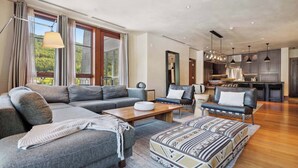 A modern living room with a sectional sofa, a coffee table, and a patterned ottoman. There is a kitchen area with pendant lighting and a dining table. - Solaris 6G East - 4 Bedroom (Vail)