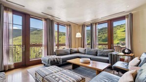 A spacious living room with a large sectional sofa, a patterned ottoman, and a wooden coffee table. Large windows offer a view of a mountainous landscape. - Solaris 6G East - 4 Bedroom (Vail)