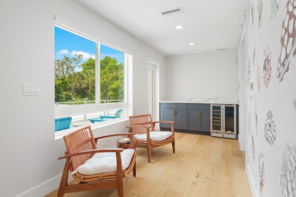 A room with a large window, wooden chairs, a small table, and a kitchen area.