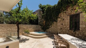 Un espace extérieur entouré de murs en pierre, comprenant un jacuzzi, un long banc et des chaises en osier.
