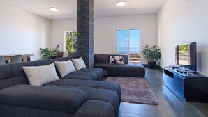 A modern living room with a sectional sofa, a stone pillar, a flat-screen TV, and a balcony with a view. - GuestReady - Spacious stay with sea views (Caniço)