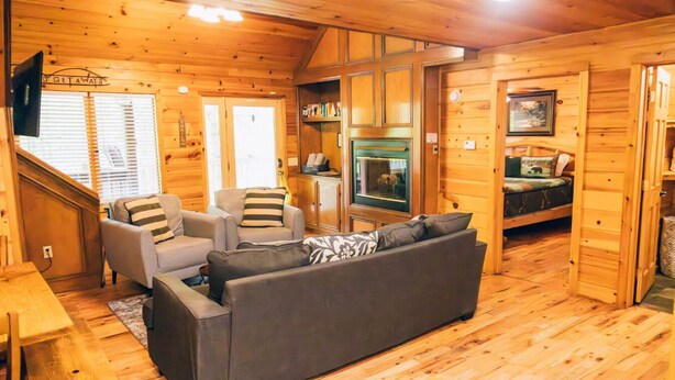 A cozy log cabin interior with a sofa, armchairs, a dining table, a kitchen area, and a television.