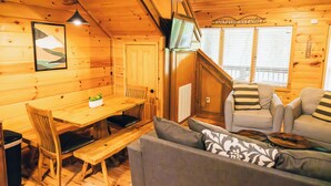 A cozy cabin interior with a wooden dining table, chairs, a sofa, and a television.