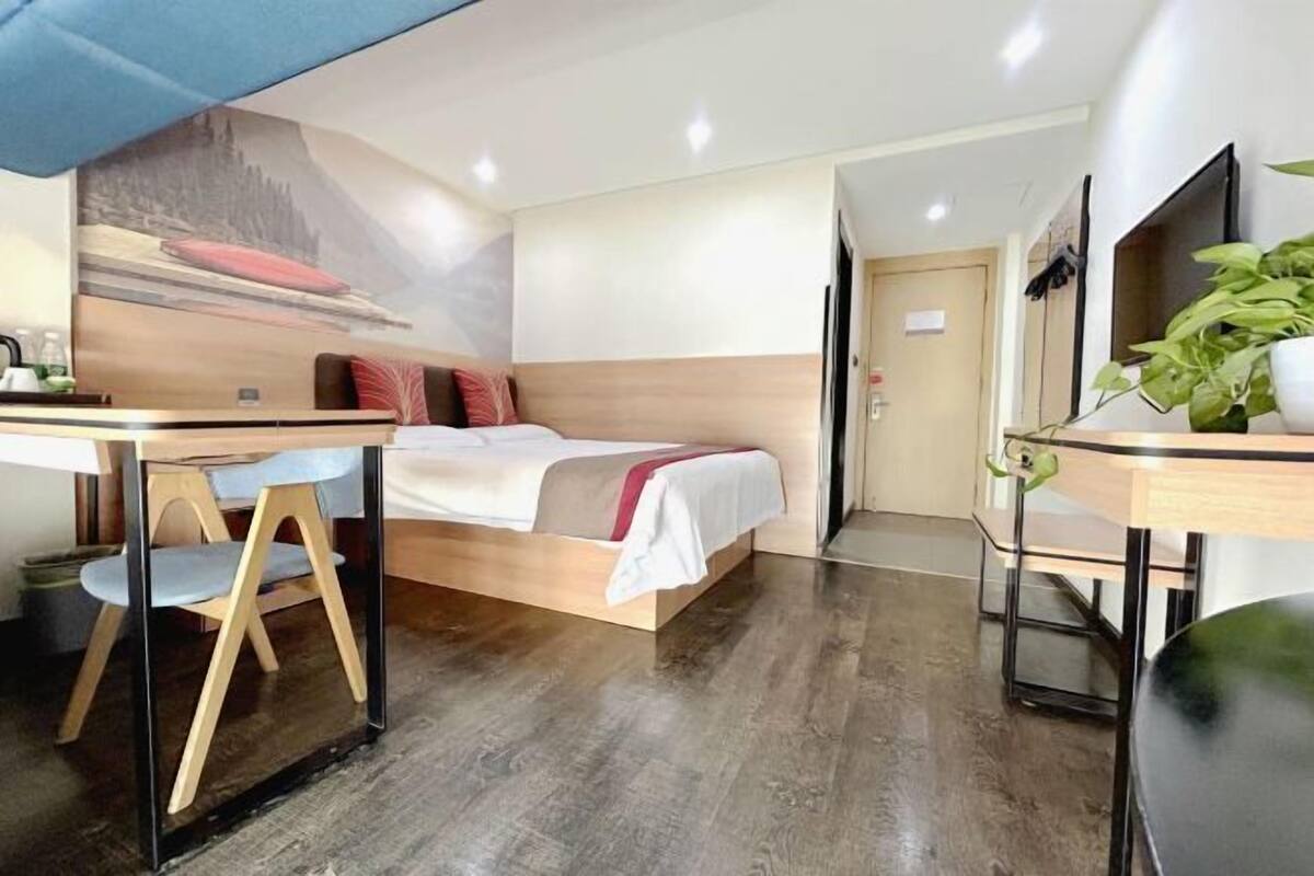 A modern hotel room with a bed, a desk, and a chair. There is a painting on the wall and a plant on the desk.