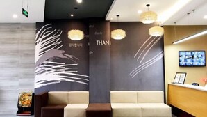 Un área de recepción moderna con dos sofás, un mostrador de madera y una pared con arte abstracto y la palabra “THANK” en varios idiomas.