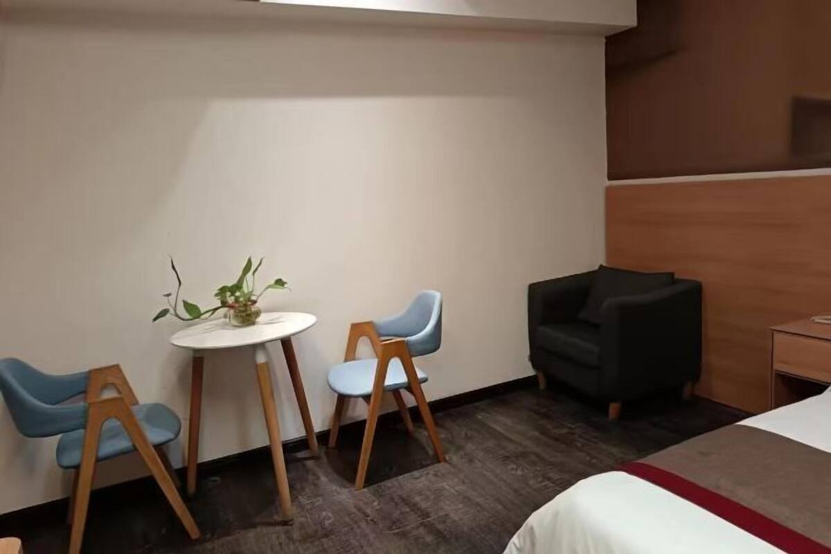 A hotel room with a bed, a small round table with chairs, a potted plant, and a dark armchair.