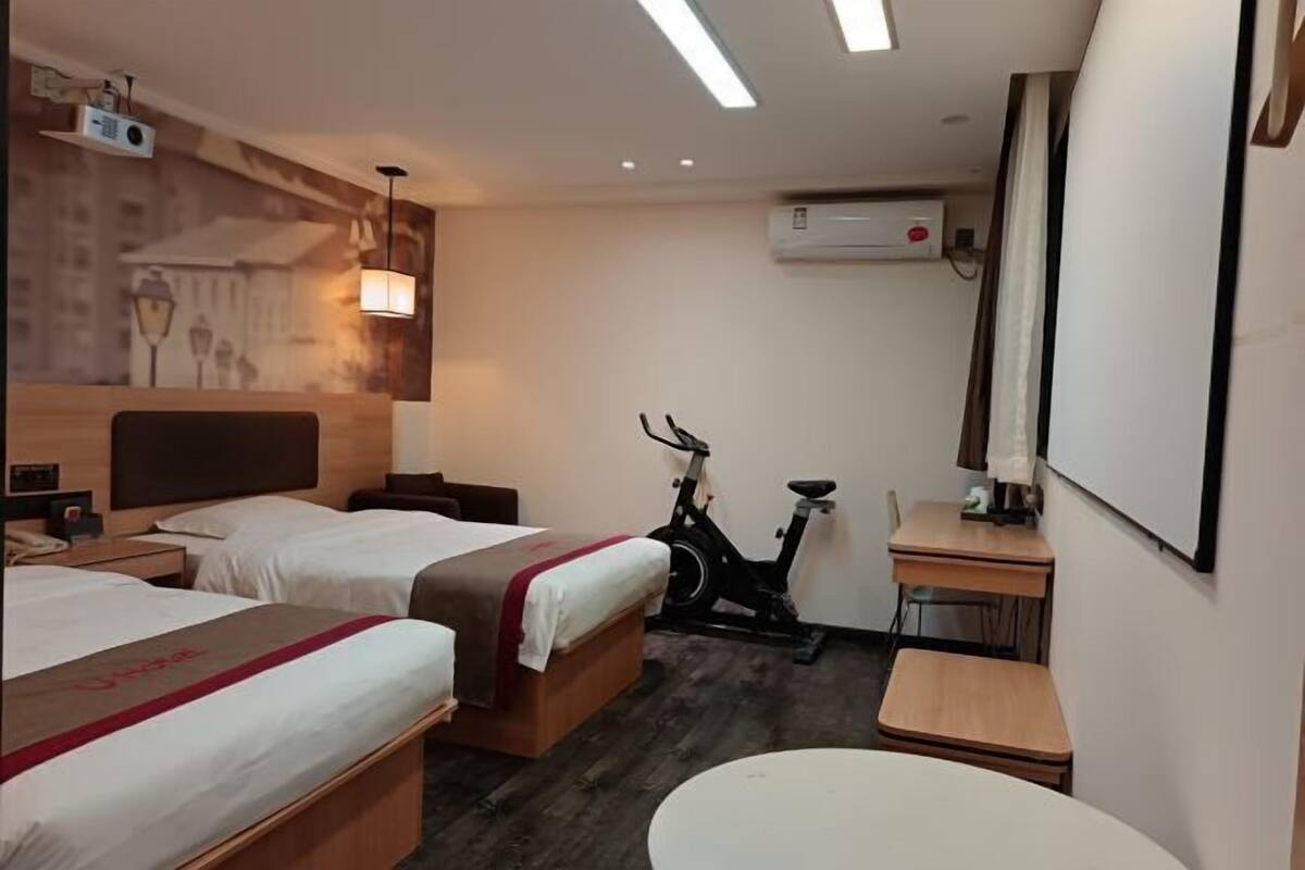 A hotel room with two beds, an exercise bike, a desk, and a wall-mounted air conditioner.