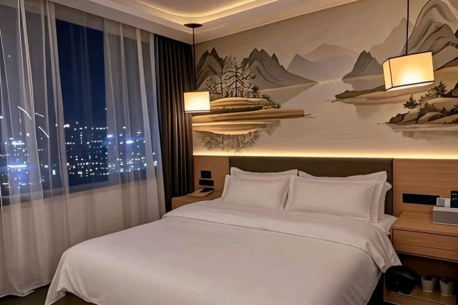 Habitación de hotel con cama, mesita de noche, lámpara, vista nocturna de la ciudad y un mural en la pared.