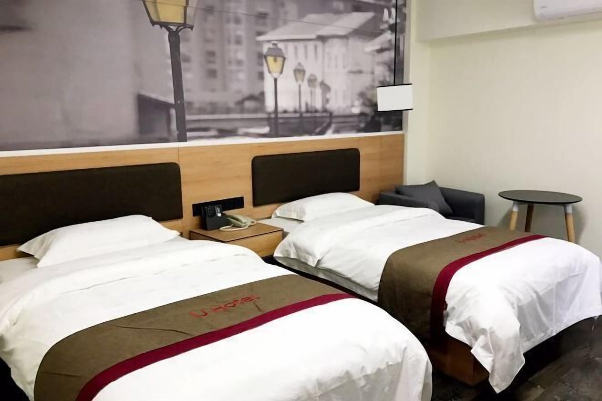 Quarto de hotel com duas camas, uma mesa de cabeceira, uma luminária e vista para a cidade.