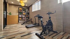 Un gimnasio con equipo de ejercicio, piso de madera, estante en la pared con objetos y una ventana.